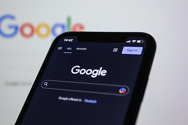 Ecr&atilde; de computador com a landing page do Google.com com um telem&oacute;vel com a mesma p&aacute;gina em dark mode &agrave; sua frente.