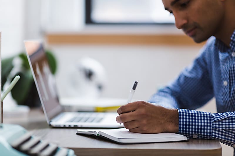 Um homem a desenhar num caderno enquanto usa um computador.