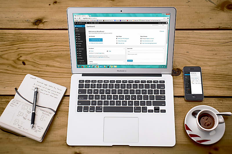 Um computador rodeado por um telem&oacute;vel, um caderno com o wireframe de um website desenhado numa das p&aacute;ginas e um caf&eacute;.